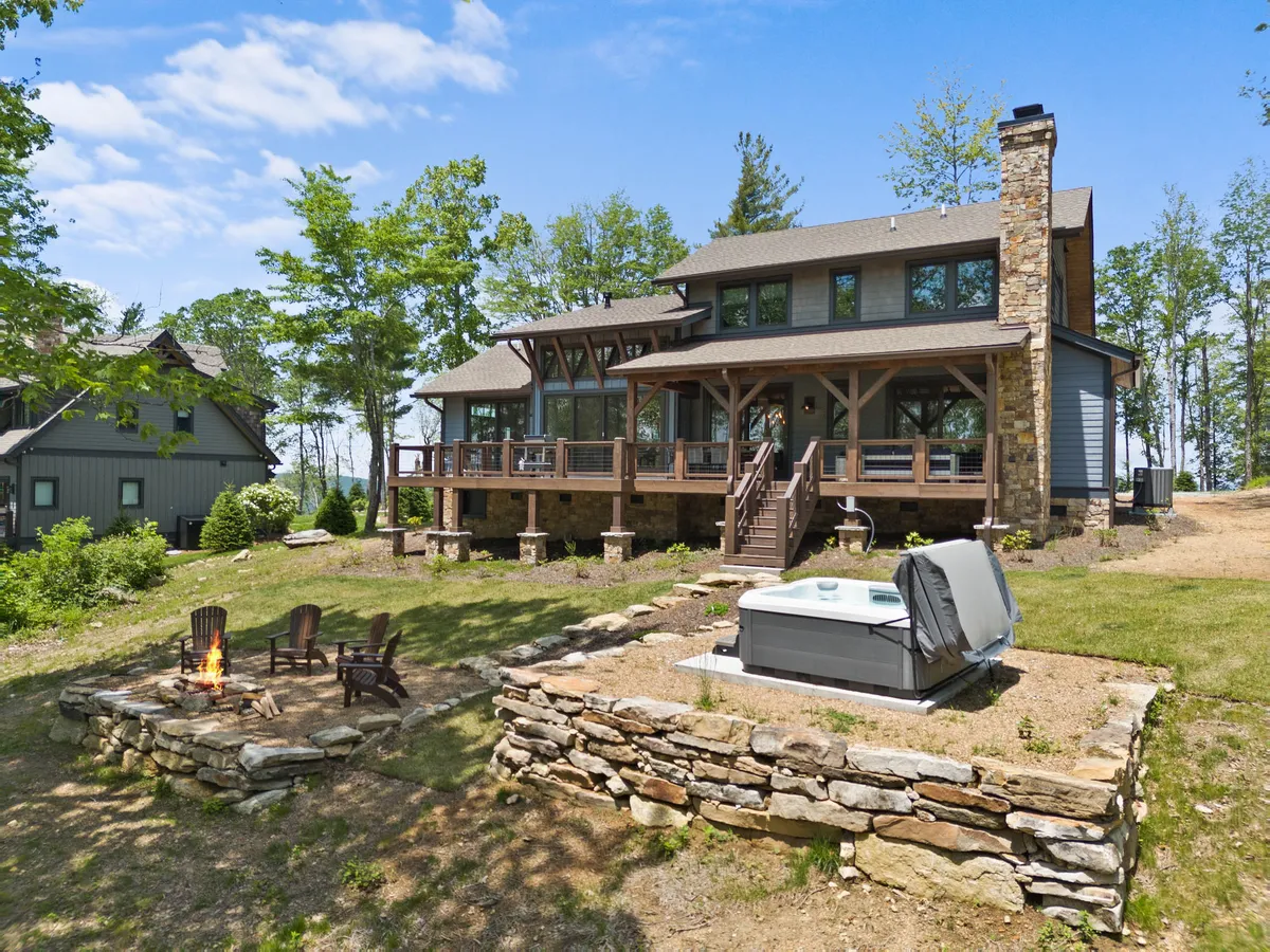 The hero image showcases a stunning, spacious cabin nestled in lush greenery, featuring a large deck with seating, a crackling fire pit, and a bubbling hot tub—all surrounded by the tranquil North Carolina mountains. The inviting outdoor area hints at unforgettable evenings watching sunsets, stargazing, and relaxing with friends and family.