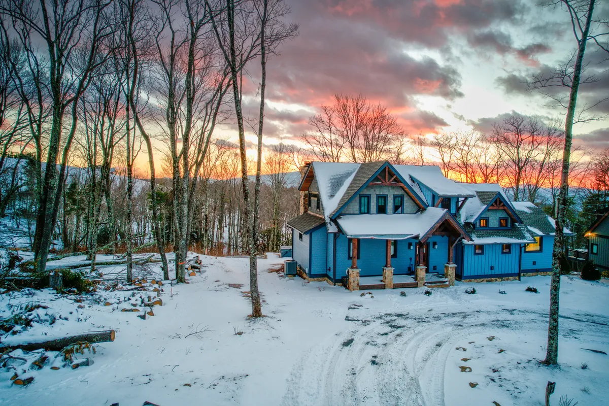 A charming blue house sits amidst a snow-covered landscape, framed by a tranquil tree line and a warm sunset sky. The inviting porch and spacious yard suggest ample room for families to play, relax, or enjoy seasonal activities like building snowmen or watching the sunset.