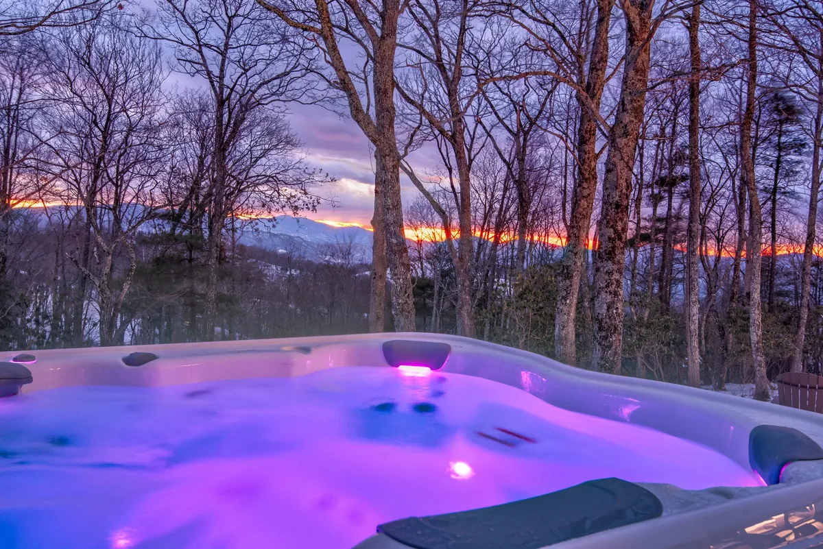 A bubbling hot tub overlooks distant mountains and silhouetted trees under vibrant sunset hues. The setting is tranquil, offering a perfect spot for relaxation or socializing after a day of adventures.