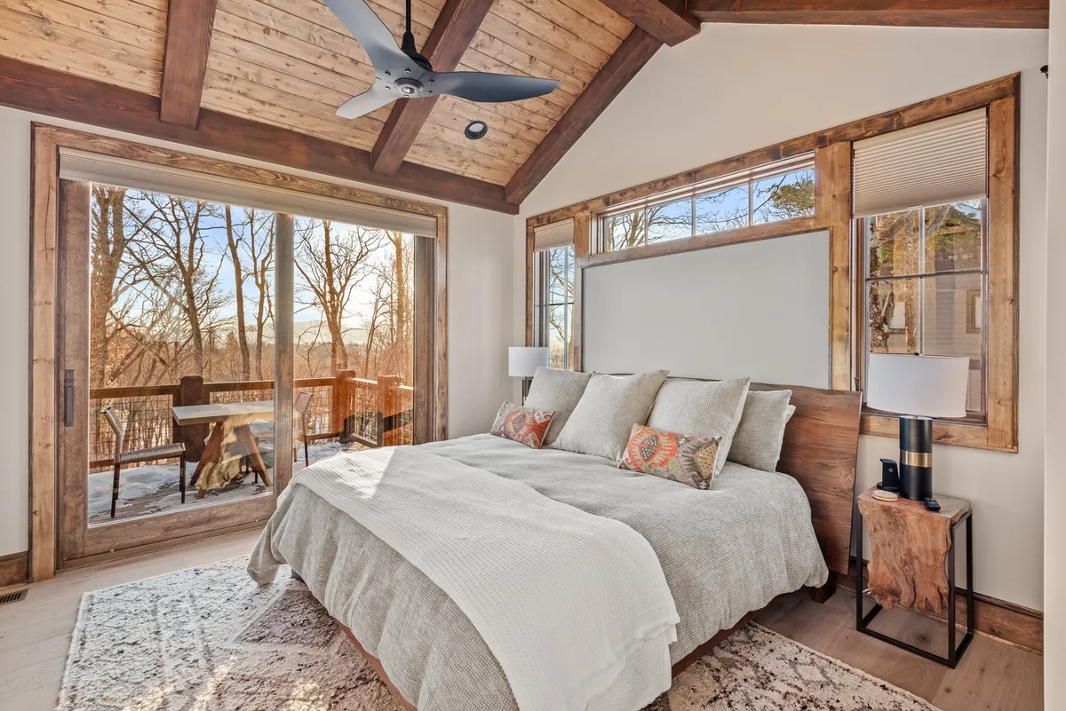 A tranquil bedroom with a cozy bed topped with textured bedding, set beside large windows that frame views of the surrounding natural landscape. Wooden accents provide warmth, and the ambiance is serene—perfect for relaxation and restful nights.