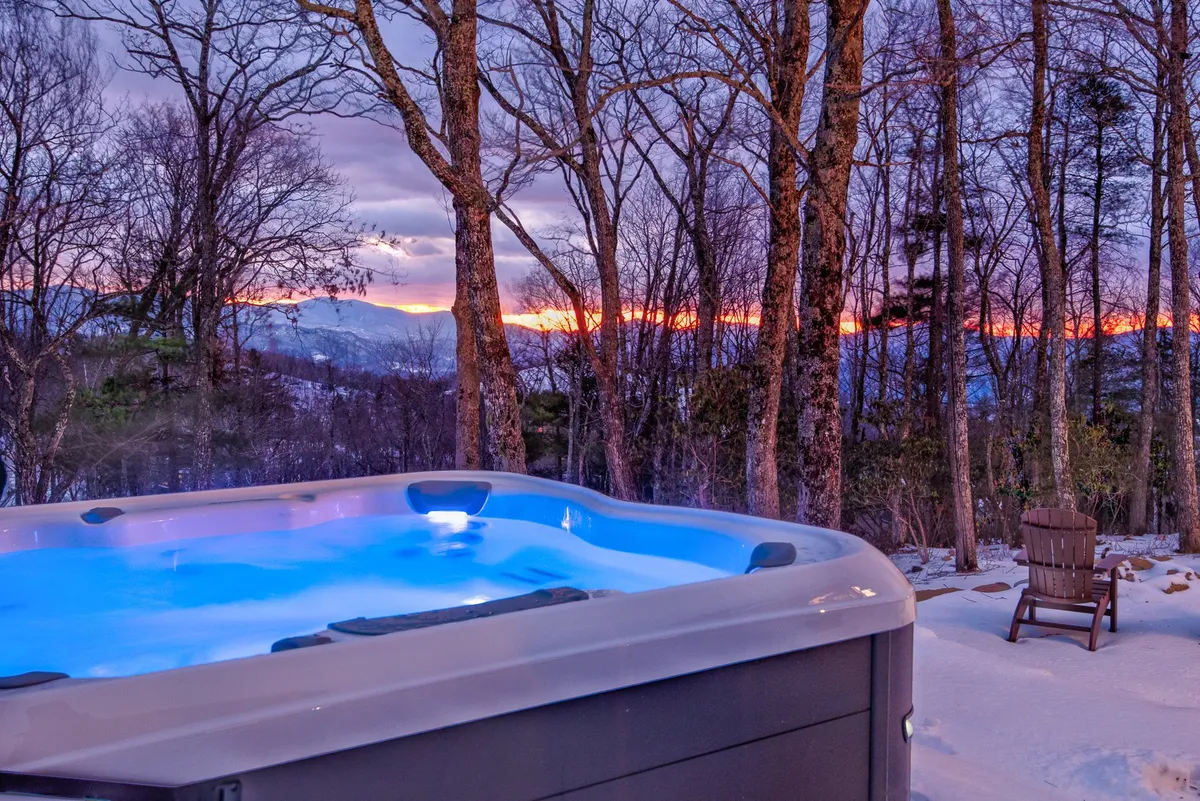 An exterior shot featuring a hot tub overlooking a sunset-lit mountain landscape, with bare trees framing a tranquil, wintry backdrop. The warm water of the hot tub contrasts with the cool tones of the snowy ground, inviting guests to relax and enjoy the view.
