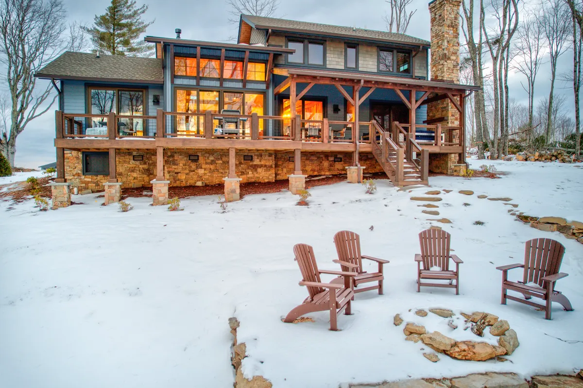 The exterior scene captures the house’s large windows aglow with warm light, a wooden deck, stone chimney, snow on the ground, and Adirondack chairs set up for relaxation. The environment is peaceful yet inviting, suggesting both comfort after outdoor activities and easy access to adventure.