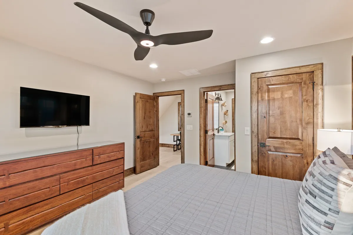 A cozy bedroom featuring a bed with textured bedding, a wooden dresser, ceiling fan, and a TV mounted on the wall. Warm wooden doors complete the inviting, contemporary look, and gentle lighting enhances the intimate feel of the space.