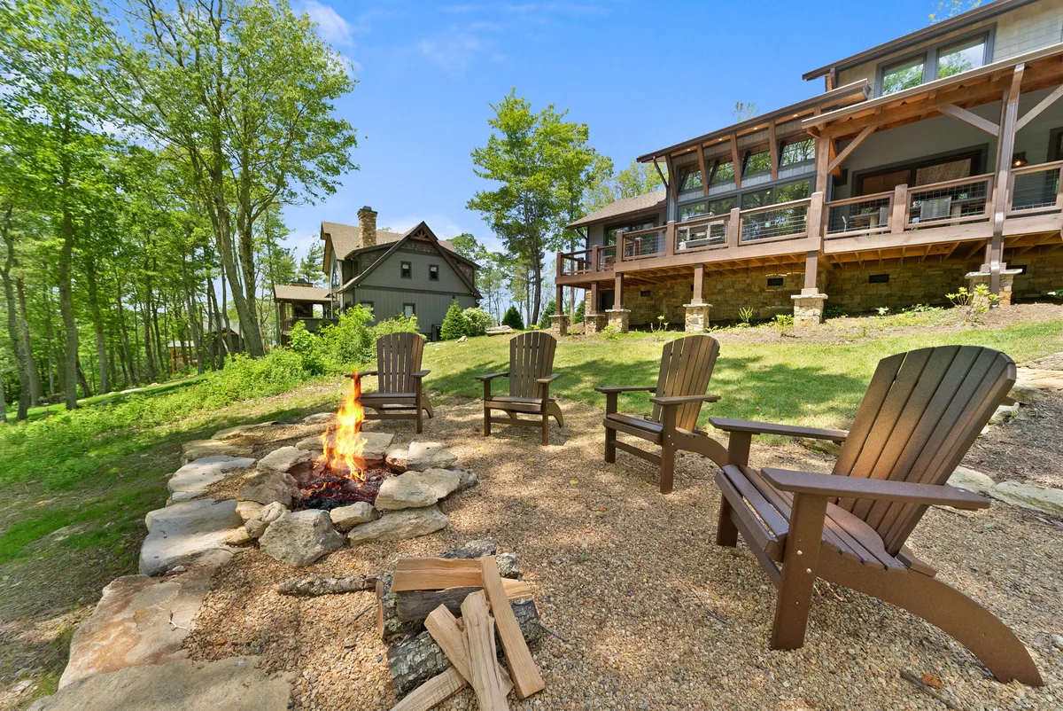 A cozy fire pit area surrounded by Adirondack chairs, set amidst tall trees with the main house visible in the background. The atmosphere is communal and inviting, with the setup ideal for relaxing outdoors and sharing stories after a day of exploring with your pet.