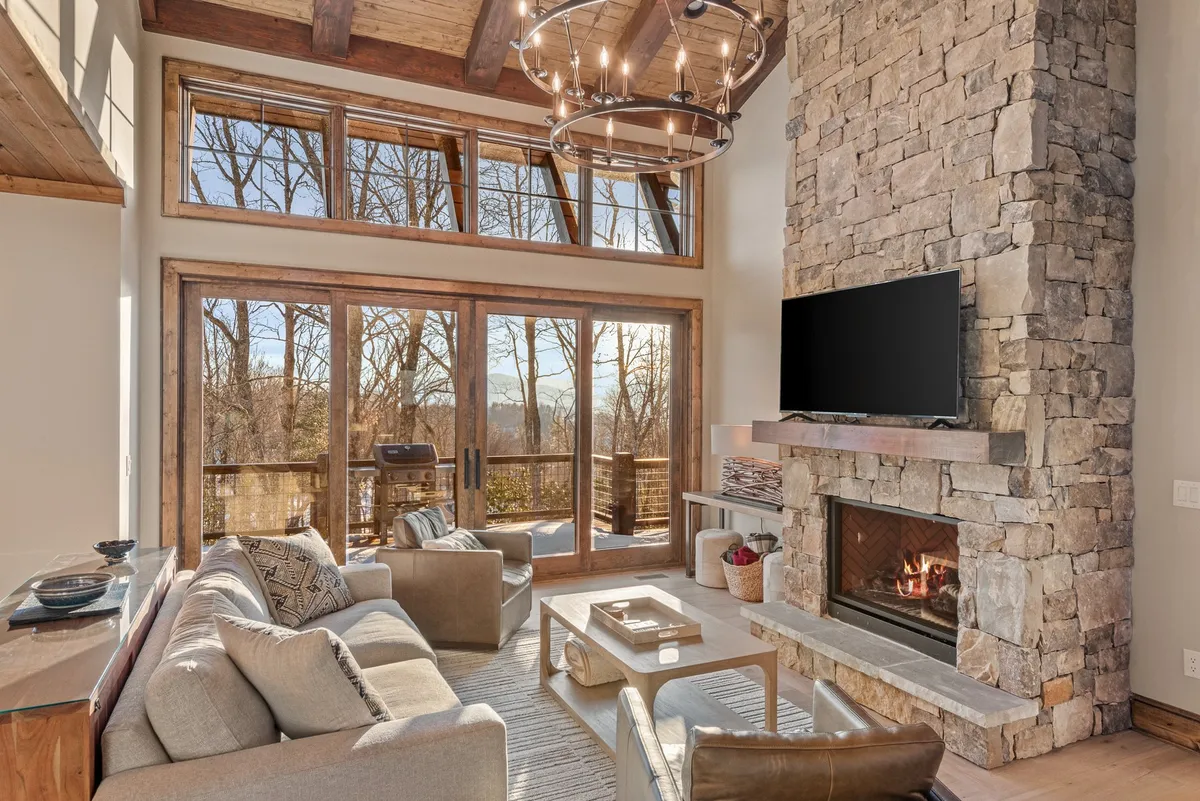 A warm, inviting great room featuring a dramatic floor-to-ceiling stone fireplace at its center, surrounded by plush sofa seating and a central coffee table. Large windows flood the space with natural light and offer scenic views, while a stylish chandelier adds a touch of elegance overhead. The arrangement hints at a cozy gathering area perfect for fire-lit evenings, relaxing, or watching TV.