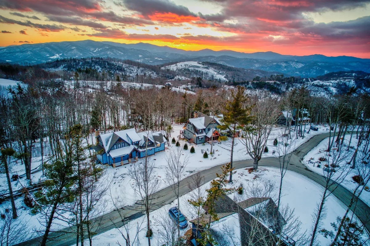 A sweeping exterior view of a snow-covered landscape at sunset, with multiple houses featuring unique architectural details. The tranquil mountains and colorful sky evoke exploration, winter sports, and the thrill of discovering new vistas.