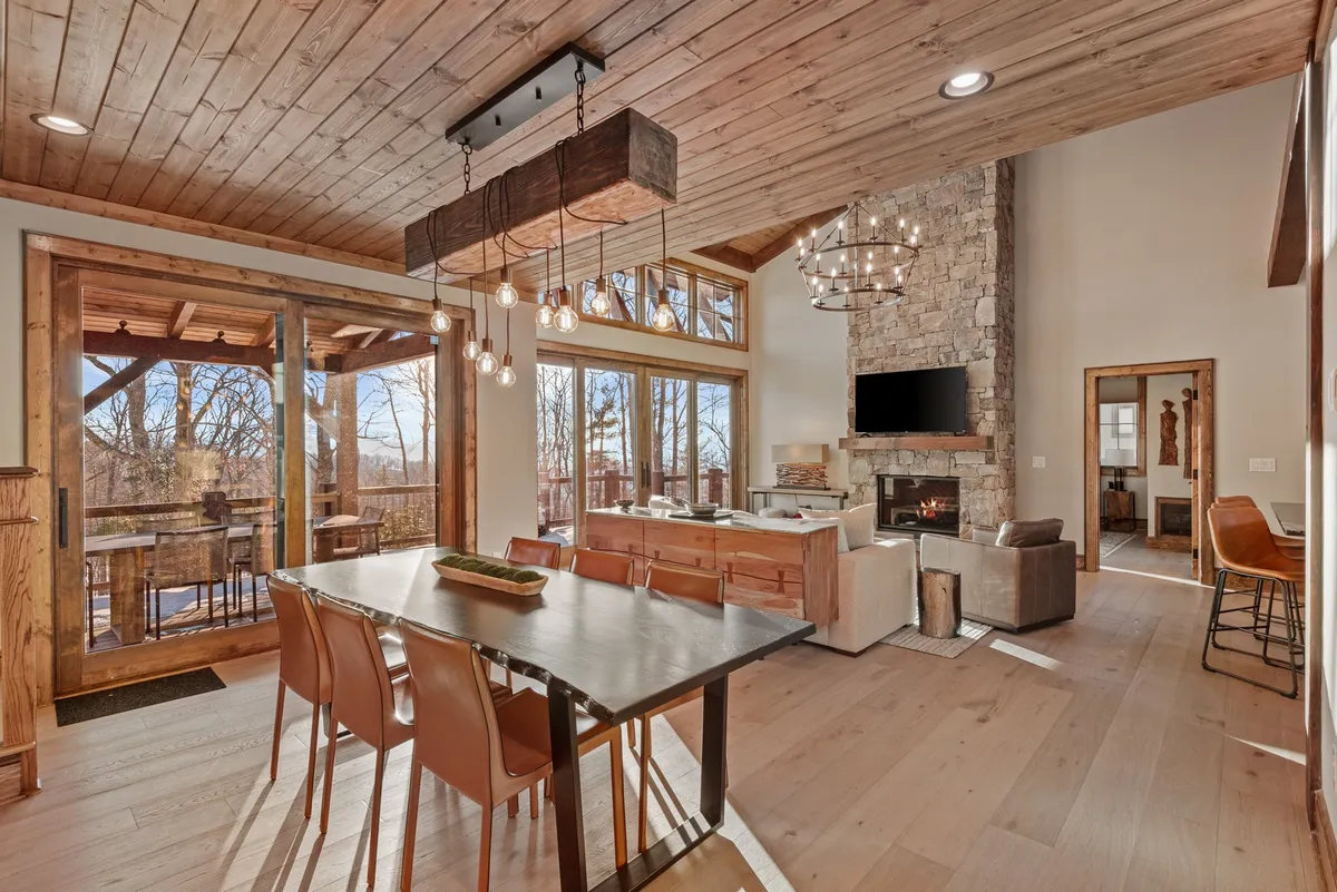 An open, expansive dining and kitchen area illuminated by ample natural light streaming through large windows. The space features a stylish dining table, a modern kitchen island with bar stools, a fireplace, and contemporary lighting. The layout encourages communal dining and effortless movement between cooking, eating, and relaxing.