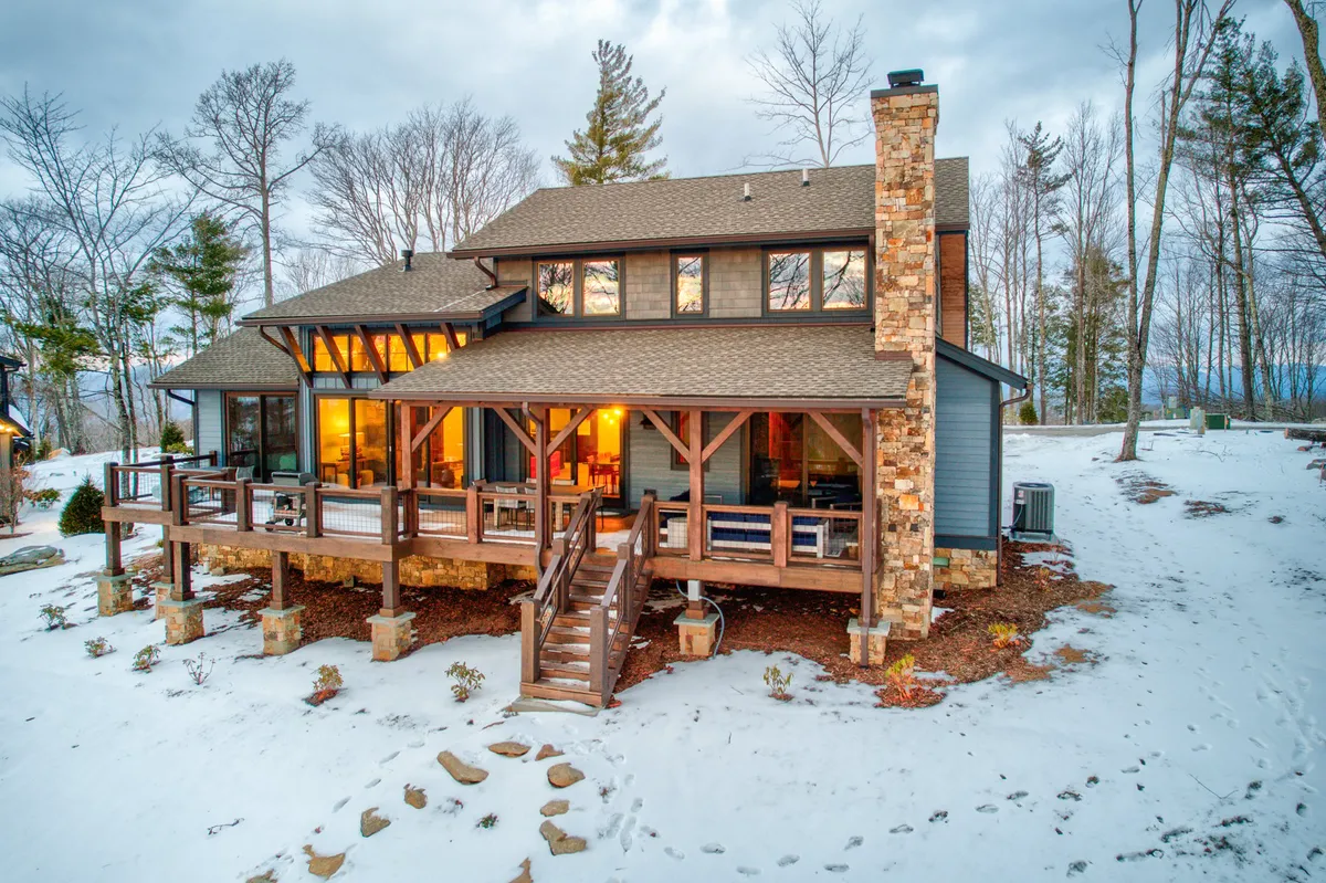 A wooden deck with large windows and a stone chimney looks out over a snow-covered yard, inviting guests to enjoy nature in all seasons. The deck is ideal for lounging with pets or watching them play in the snow.