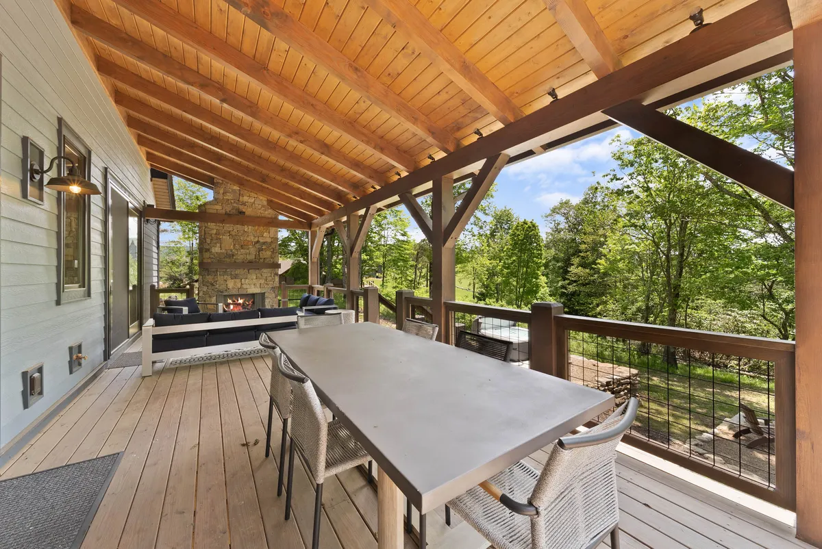 A spacious, partially covered deck featuring a large outdoor dining table and a comfortable seating area with a modern gas log fireplace. Lush green trees form a tranquil backdrop, setting the stage for lively gatherings or peaceful sunset dinners.